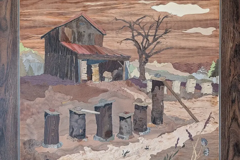 Marqueterie de bois représentant un rucher en hiver : une rangée de ruches sombres sur la neige devant une cabane et un arbre dénudé